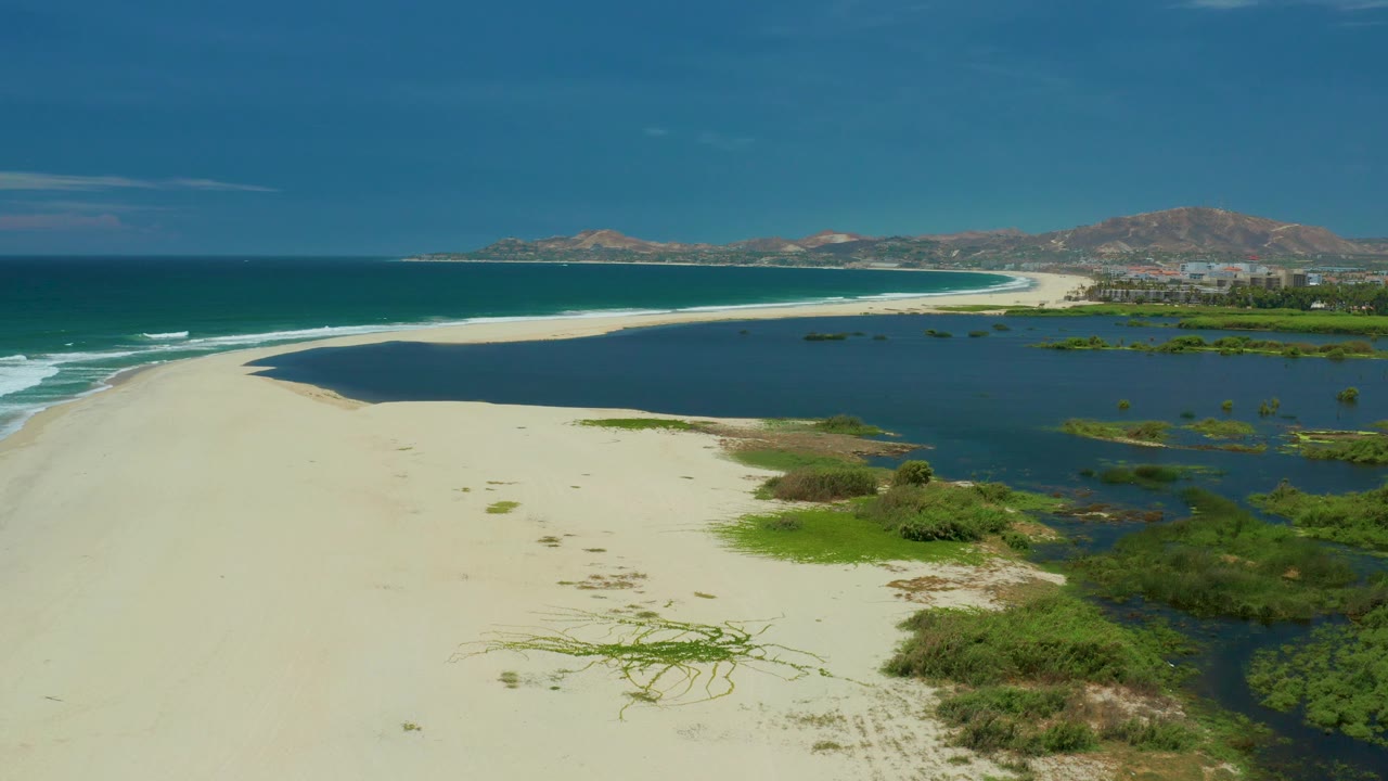 San Jose Del Cabo beach aerial with a pale sand curve leading toward distant hills, white wave lines on the ocean side, dark lagoon water, green bushes, and no visible people.