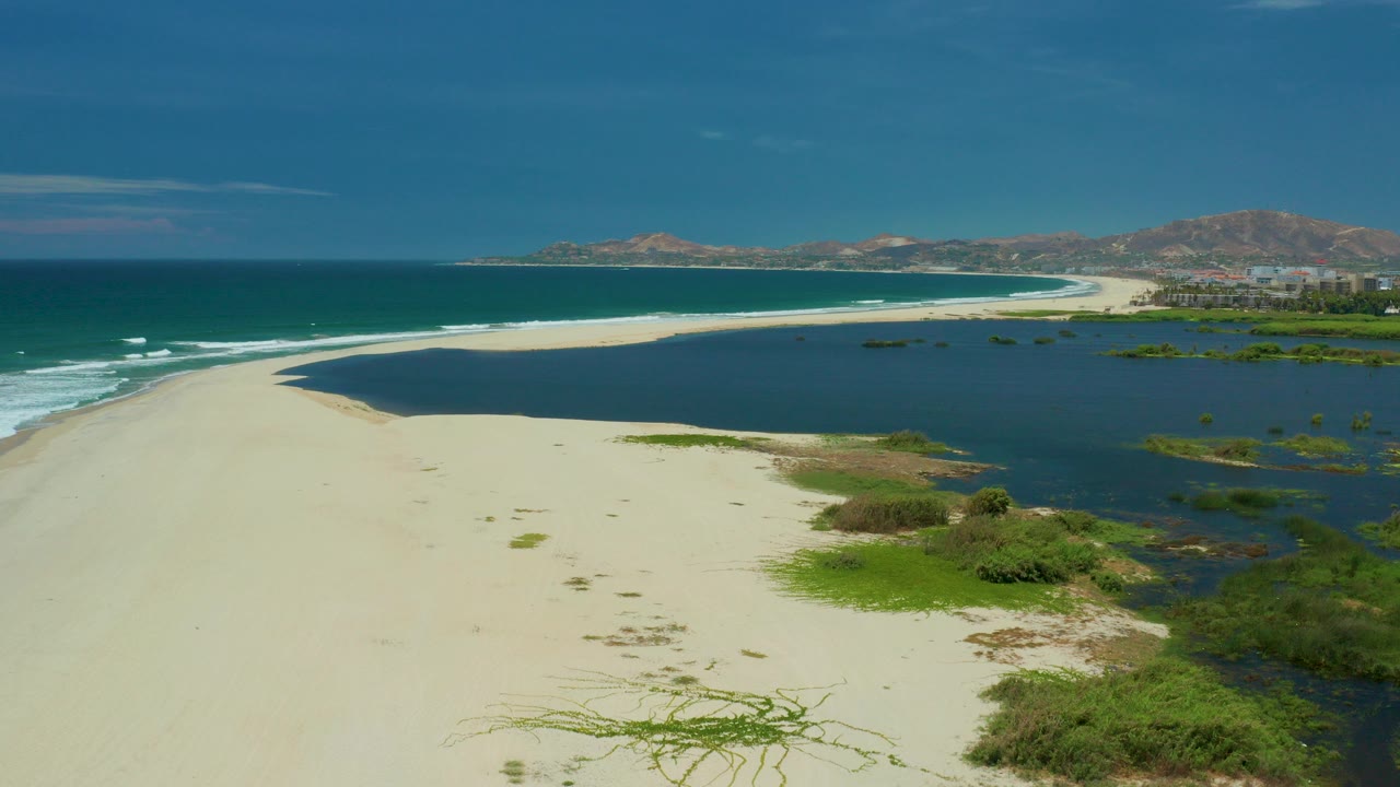 A wide aerial frame over San Jose Del Cabo, Mexico shows white sand curving between blue ocean on the left and dark wetland water on the right, with distant hills and no visible people.