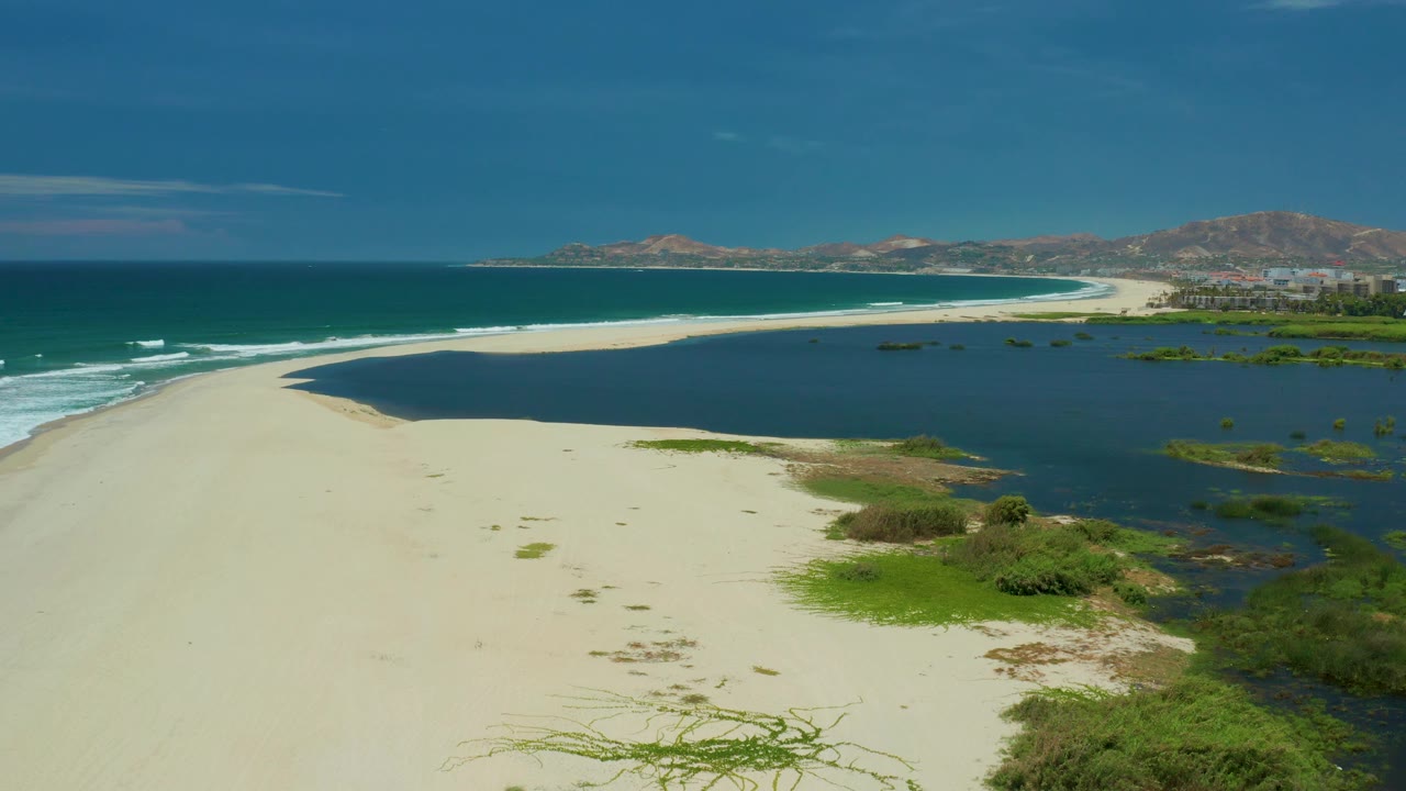 A bright aerial view in San Jose Del Cabo, Mexico shows the sandy beach running toward distant hotel buildings, with blue ocean left, dark wetland water right, and no visible people.