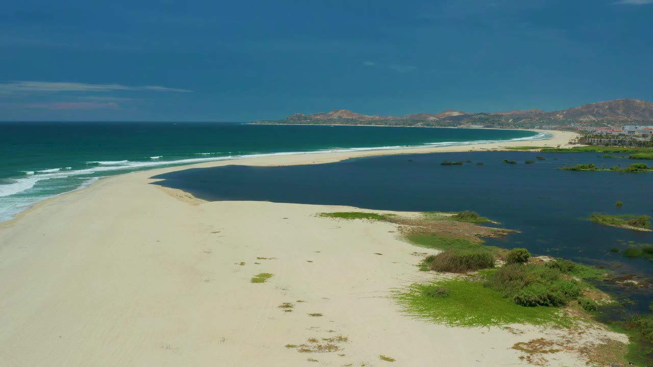 A wide drone frame in San Jose del Cabo shows the sandbar curving between white surf and the darker lagoon, with green scrub in the lower right and no visible people.
