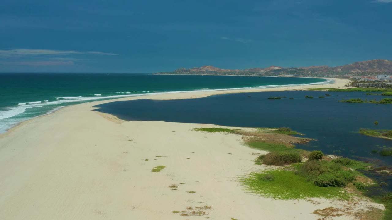 San Jose del Cabo coastline from above, with empty pale sand in front, dark lagoon water to the right, breaking surf to the left, distant shoreline, and no visible people.