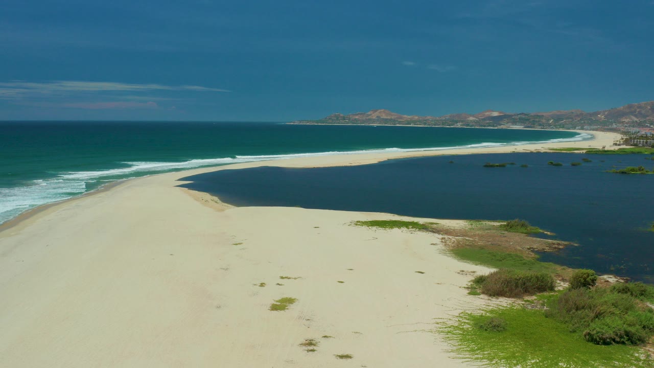 Wide aerial frame of the San Jose Del Cabo coast with pale sand in front, surf breaking along the left edge, dark lagoon water to the right, distant hills on the horizon, and no visible people.