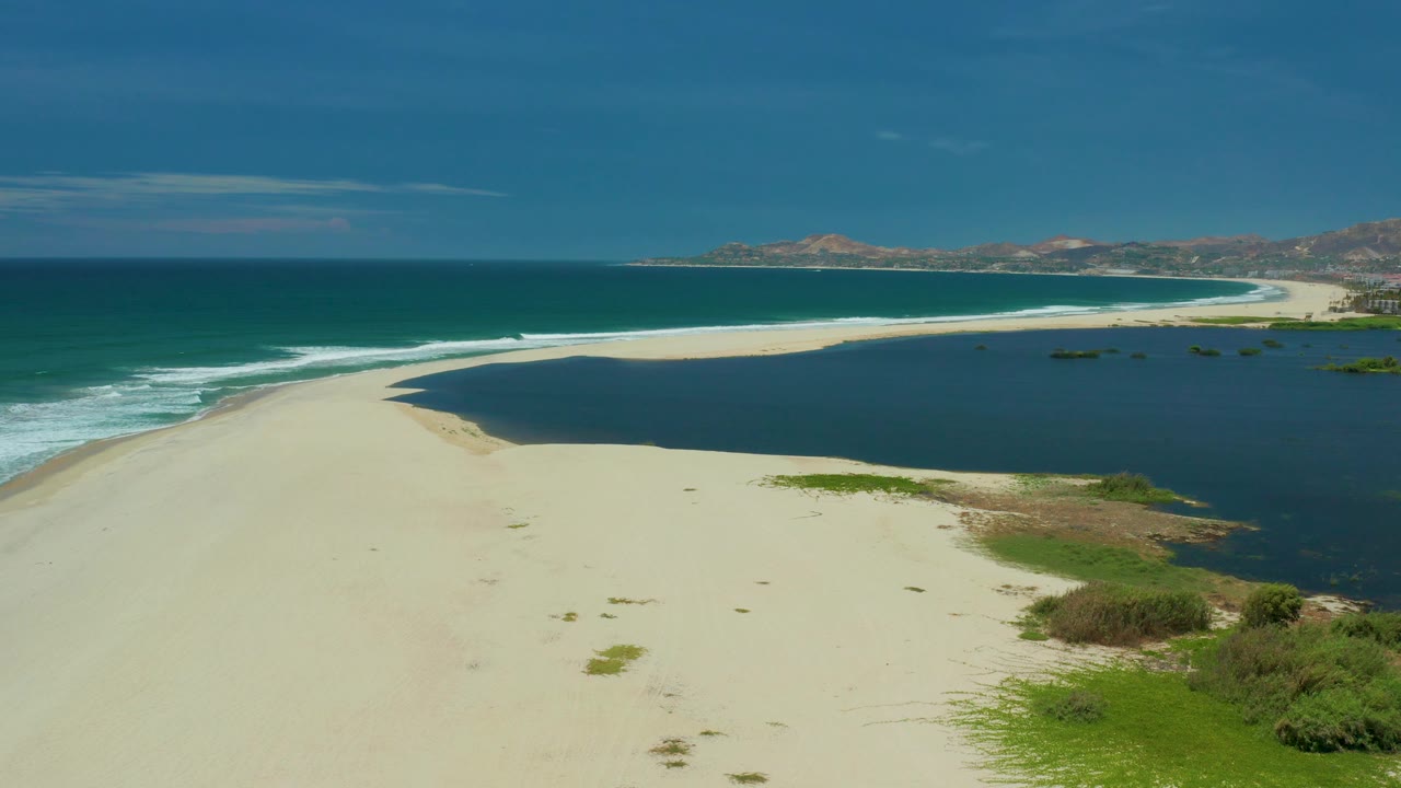 San Jose Del Cabo aerial frame showing an open pale beach, a curved edge of lagoon water, surf breaking along the ocean side, low vegetation at the lower right, and no visible people.