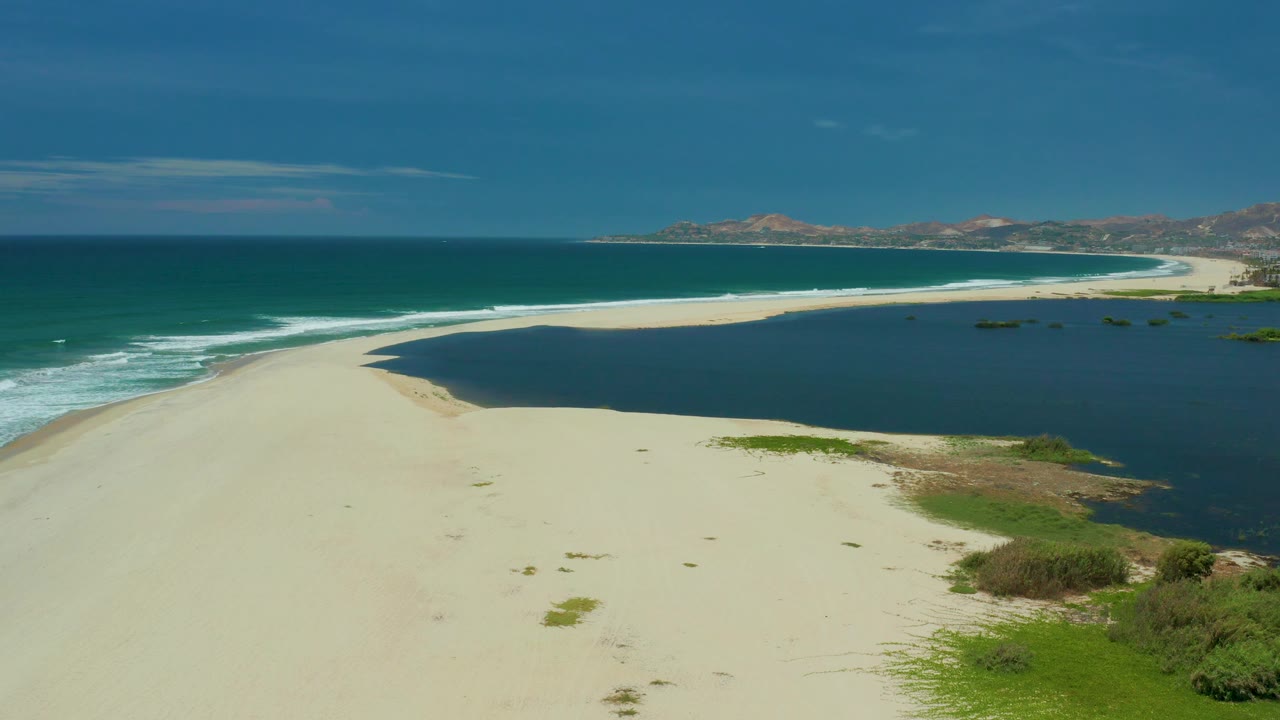 Wide San Jose Del Cabo coast view with wind-textured ocean waves on the left, still darker water on the right, a broad pale sand foreground, distant hills, and no visible people.