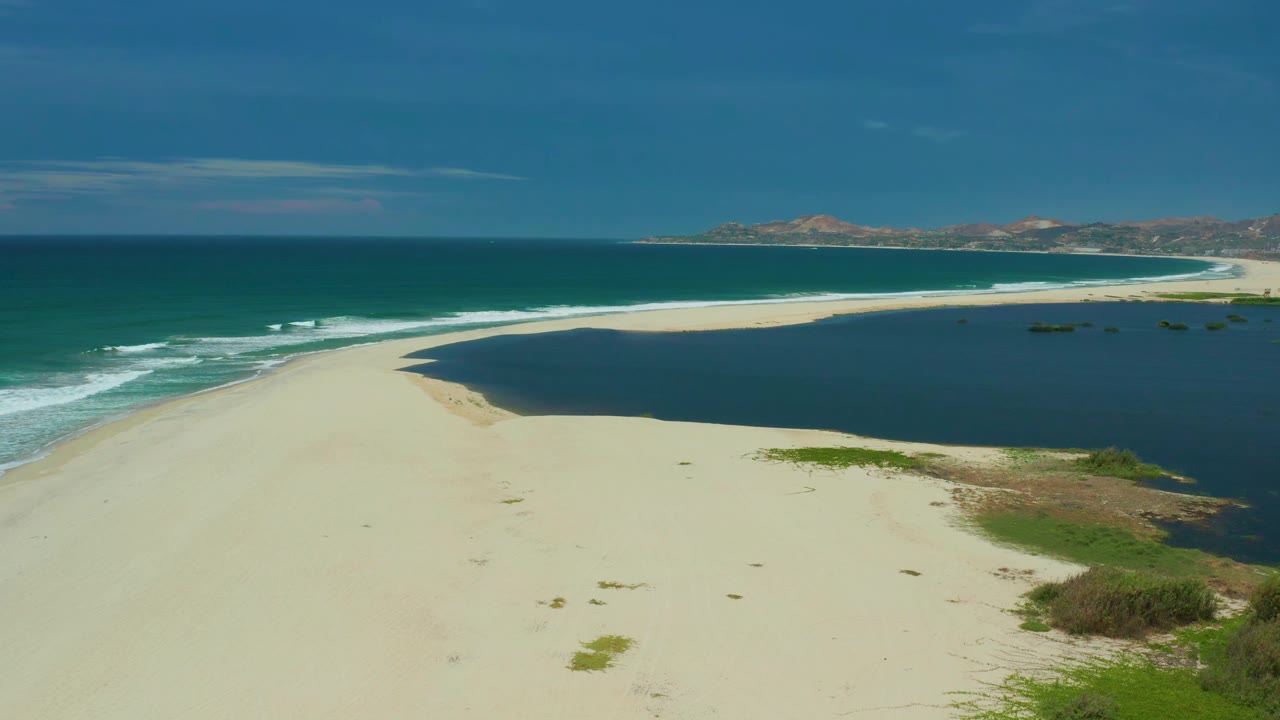 A wide San Jose Del Cabo coastline view shows the sandbar stretching across the frame, breaking waves along the left edge, dark lagoon water to the right, and no visible people.