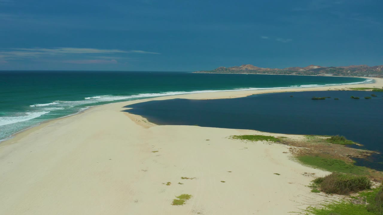The San Jose Del Cabo beach remains empty as pale sand fills the foreground, white surf lines the left shoreline, dark lagoon water fills the right side, and no visible people appear.