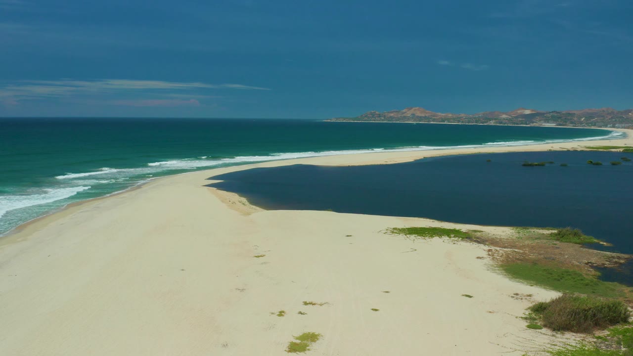 A high coastal view in San Jose Del Cabo shows waves rolling in from the left, a broad pale beach in the lower frame, the lagoon curving to the right, and no visible people.