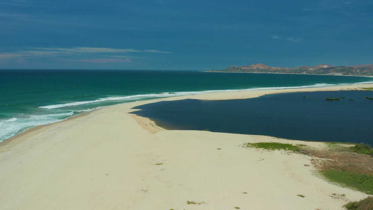 Aerial view in San Jose Del Cabo showing pale sand between green-blue surf and a dark lagoon, with distant hills under a blue sky and no visible people.