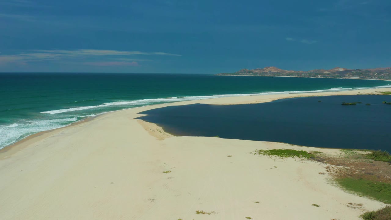 Wide aerial frame of San Jose Del Cabo coastline with white wave bands beside a sandy spit, dark lagoon water to the right, and no visible people.