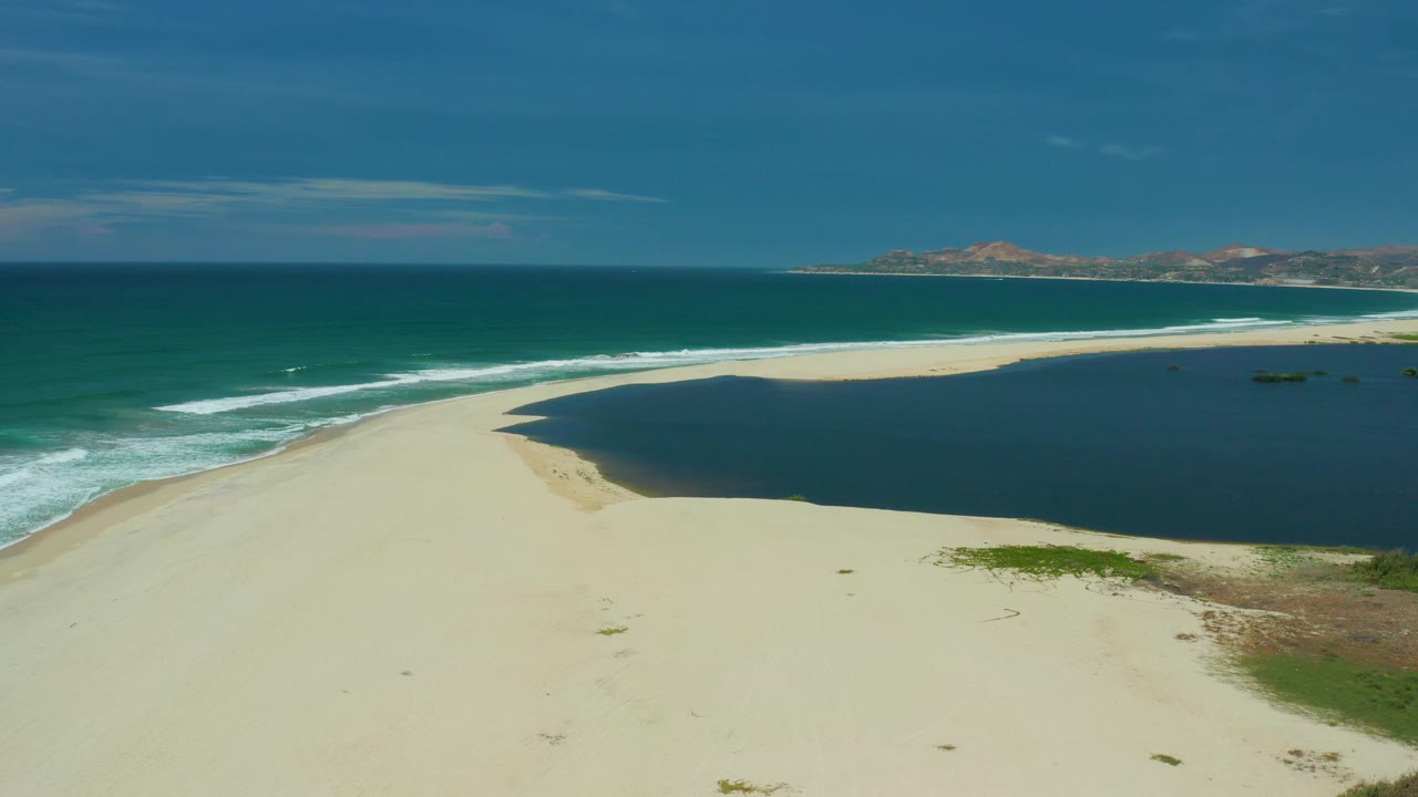 Empty sandbar in San Jose Del Cabo curves between green ocean water and a darker lagoon, with low hills along the horizon and no visible people.