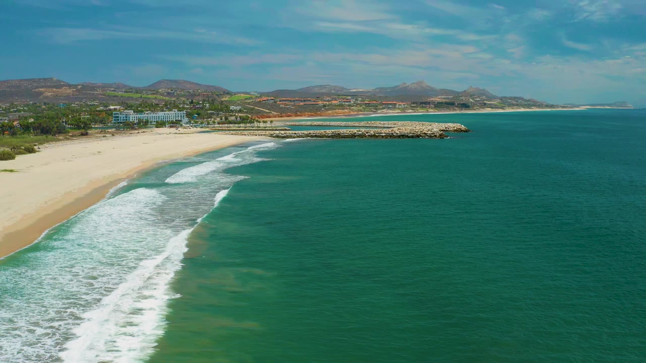 Aerial view in San Jose Del Cabo, Mexico, with white surf reaching a pale sandy beach, open blue green water to the right, trees and hills inland, and no visible people.