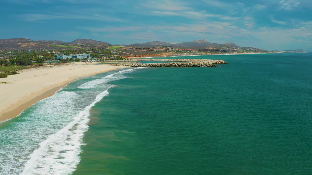 San Jose Del Cabo aerial beach frame showing foamy waves pushing farther up the shore, open water dominating the right side, a stone breakwater, inland buildings, and no visible people.