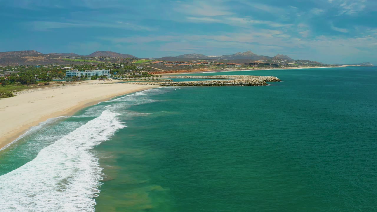 San Jose Del Cabo coastline from above with the surf band slightly farther down the beach, a low rock breakwater near the center, green shoreline vegetation, and no visible people.