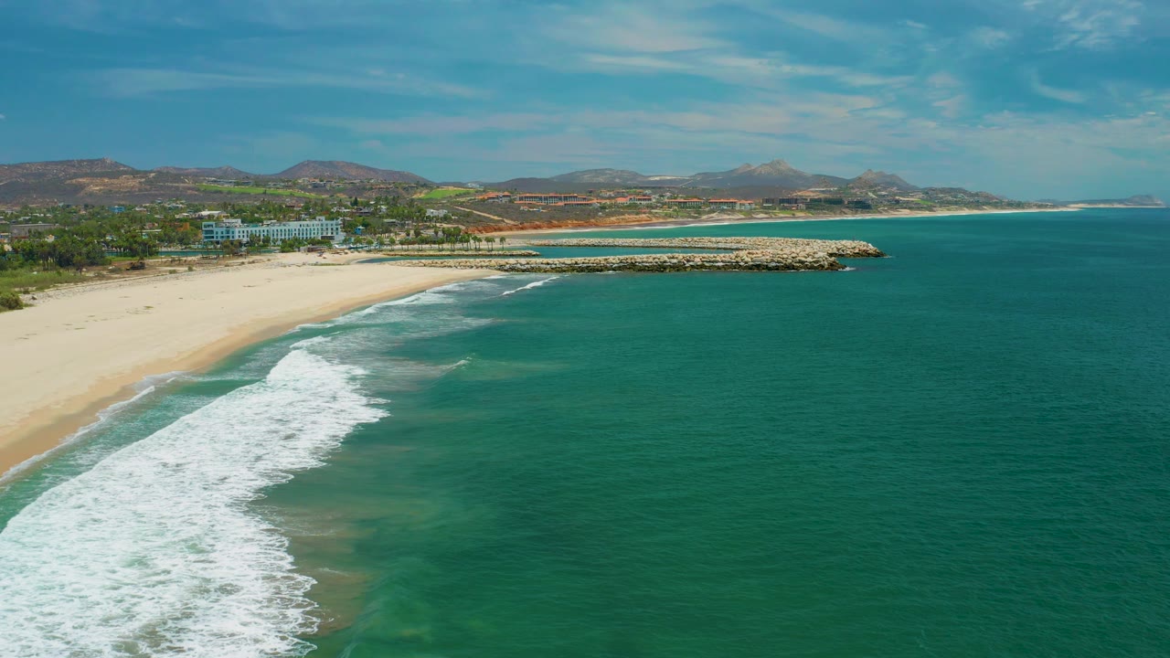 San Jose Del Cabo aerial frame with the white surf spread lower in frame, pale sand along the left, the beachfront building set back from shore, hills on the horizon, and no visible people.