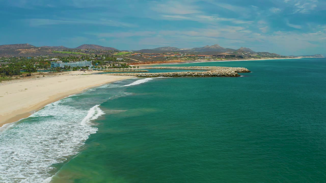San Jose Del Cabo shoreline seen from above, with the surf line slightly farther along the sand, the rock breakwater near center, low coastal buildings behind it, and no visible people.