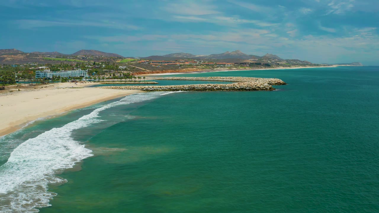 Wide aerial frame from San Jose Del Cabo, Mexico, showing a curling white wave along the left beach edge, green-blue water across the foreground, a rock breakwater, low coastal buildings, distant hills, and no visible people.
