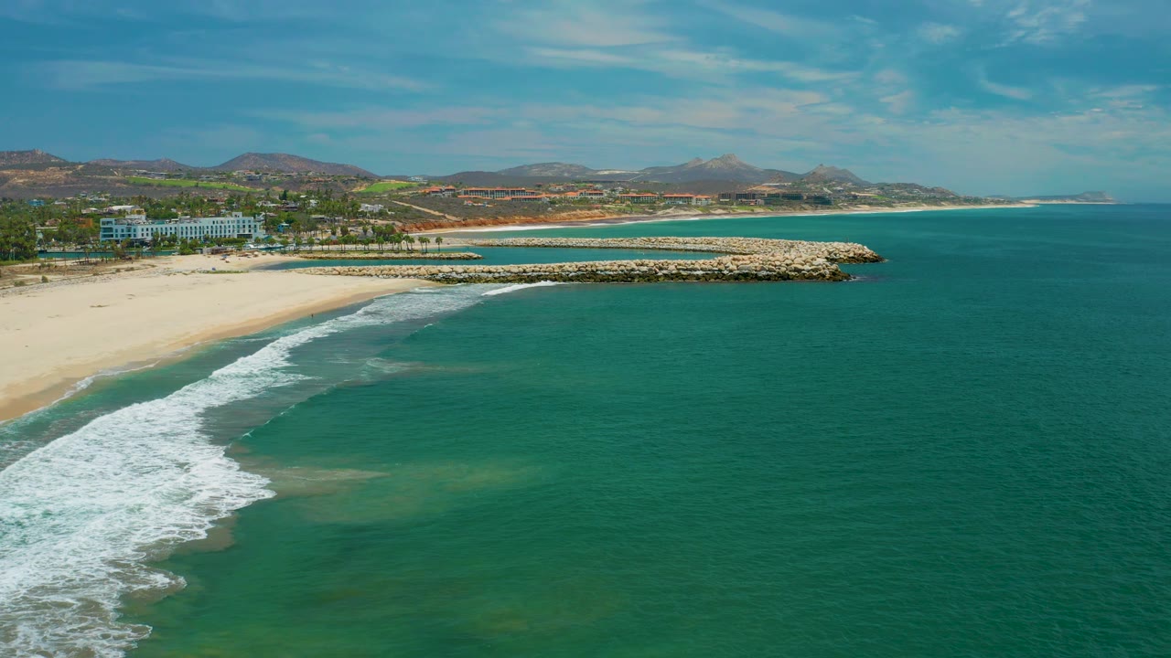 Aerial coastal view in San Jose Del Cabo, Mexico, with blue-green water taking most of the frame, foamy surf tracing the beach, a long rock structure projecting from shore, low hills in back, and no visible people.
