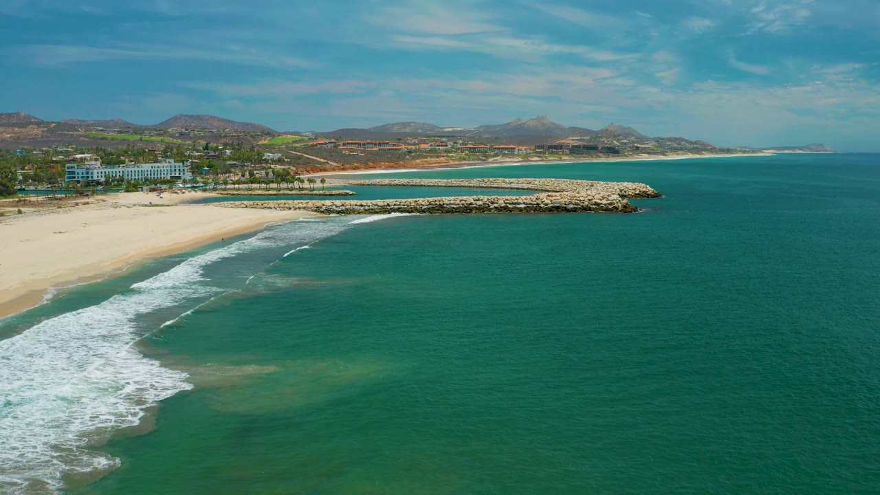 San Jose Del Cabo, Mexico, appears from above with white surf entering the lower left, a pale beach and rock jetty across the middle, teal water to the right, low hills beyond the shore, and no visible people.