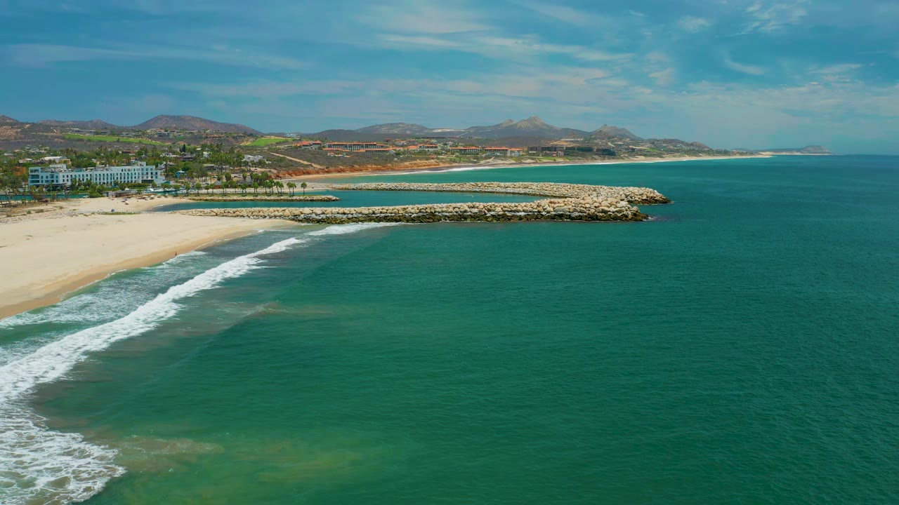 Aerial frame over San Jose Del Cabo with the surf line slightly farther along the beach, green-blue water across the foreground, a rock breakwater crossing the midground, hills beyond, and no visible people.