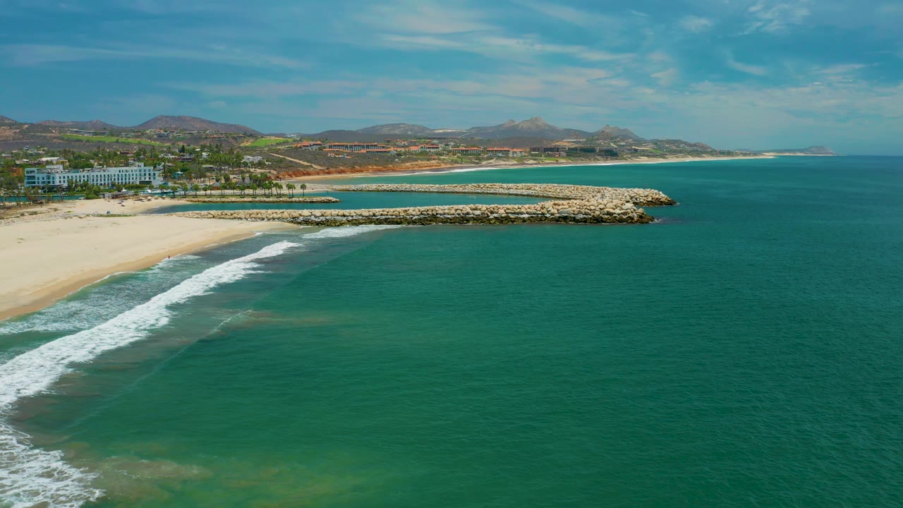 Aerial frame over San Jose Del Cabo with more open water on the right, white surf tracking diagonally from the beach, the rock breakwater centered near shore, background hills, and no visible people.
