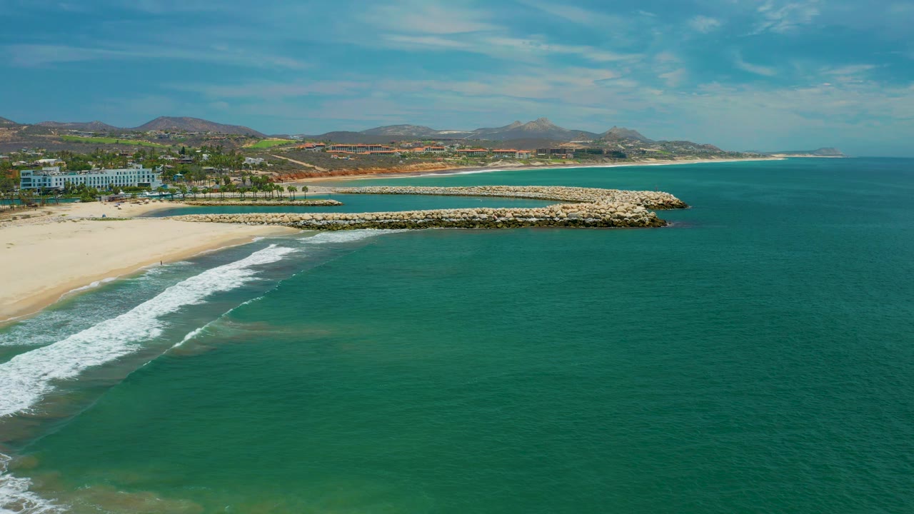Aerial frame over San Jose Del Cabo where the surf forms a longer white diagonal, the sandy beach stays at left, the breakwater spans the midground, green-blue ocean fills the right side, and no visible people appear.