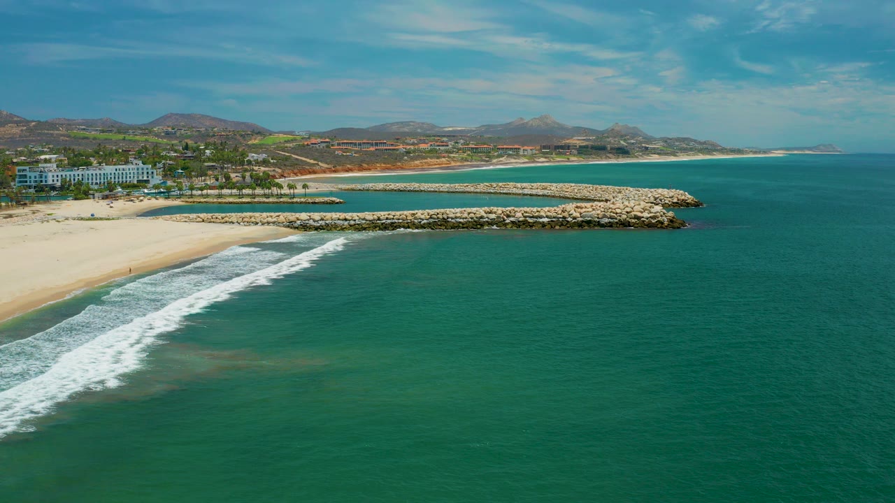 Wide aerial frame of San Jose del Cabo, Mexico with turquoise ocean in front, white surf reaching pale sand, curved rock structures offshore, coastal buildings, palms, and no visible people.
