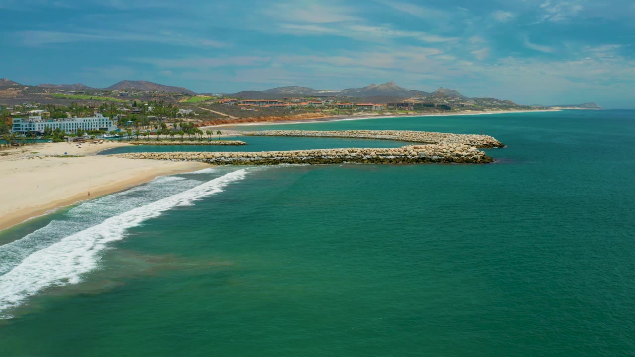 San Jose del Cabo, Mexico shoreline from above with the breaking wave shifted farther across the lower left water, rock breakwaters forming a curved edge near shore, buildings and palms to the left, and no visible people.