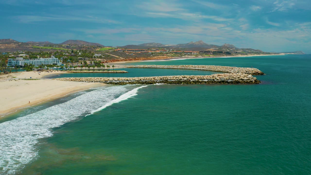 Wide aerial view of a San Jose Del Cabo beach with turquoise water filling the foreground, white surf on sand, rock breakwaters, distant hills, resort buildings at left, and no visible people.
