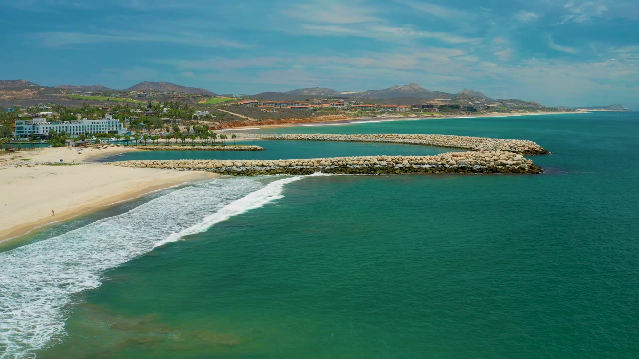 Elevated San Jose Del Cabo coastline view with more open turquoise water to the right, surf breaking along the lower beach, two rock breakwaters, resort buildings at left, and no visible people.