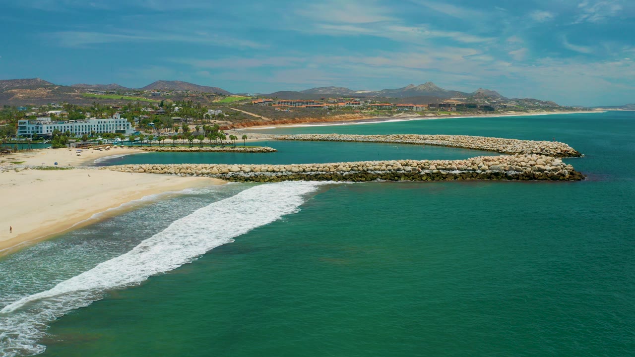 Opening aerial frame in San Jose del Cabo, Mexico, showing turquoise water in the foreground, a white surf band at the sand, a rock breakwater across the middle, resort buildings to the left, hills beyond, and no visible people.