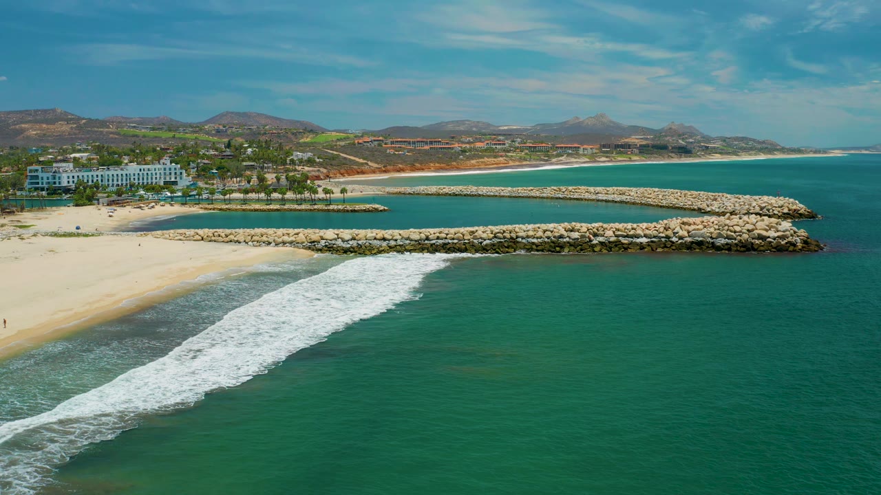 San Jose del Cabo, Mexico aerial view with the white wave line stretching diagonally from the lower left, pale sand behind it, a stone breakwater running horizontally, resort buildings near the left beach edge, distant hills, and no visible people.