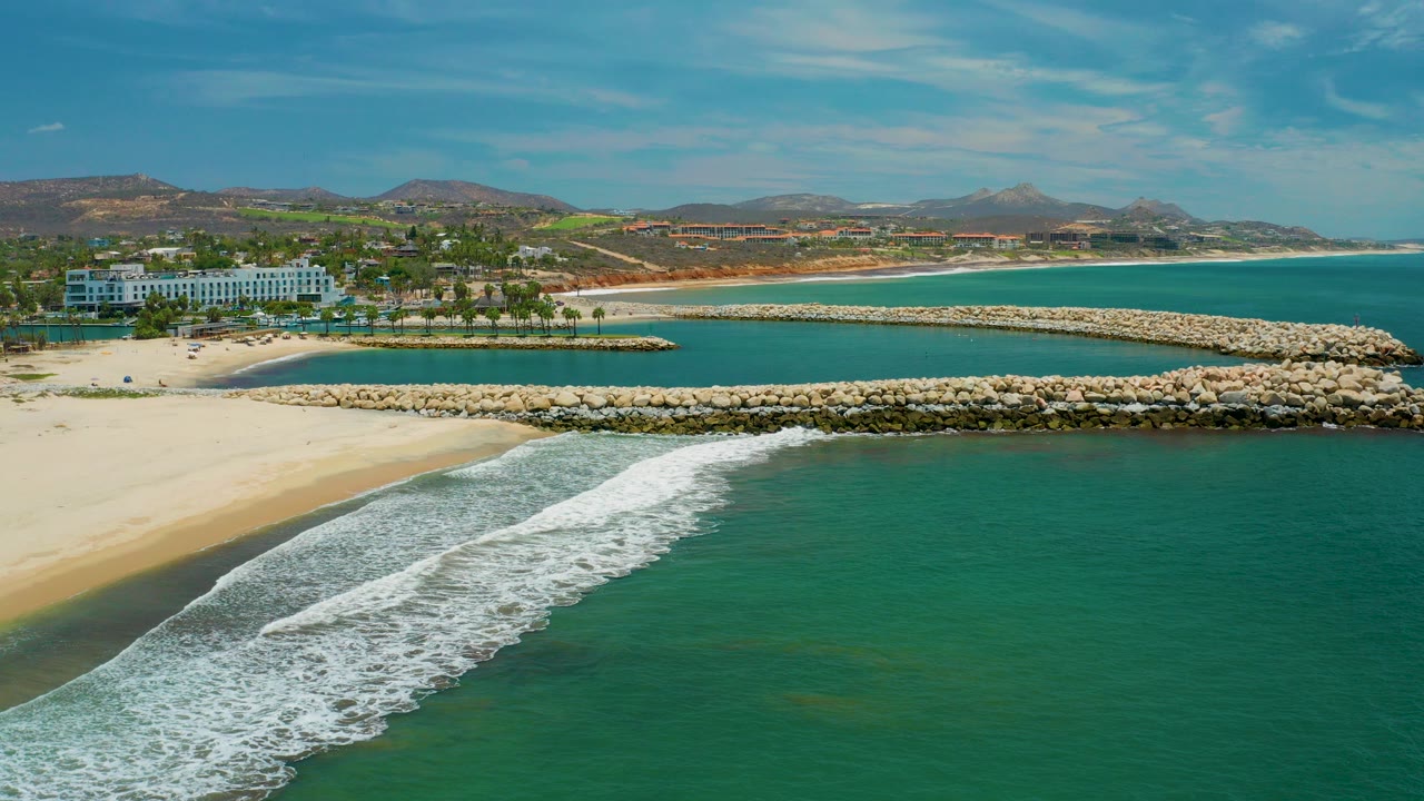 Aerial frame over San Jose Del Cabo showing white surf crossing green-blue water toward pale sand, a rock breakwater, distant resort buildings, low hills, and no visible people.