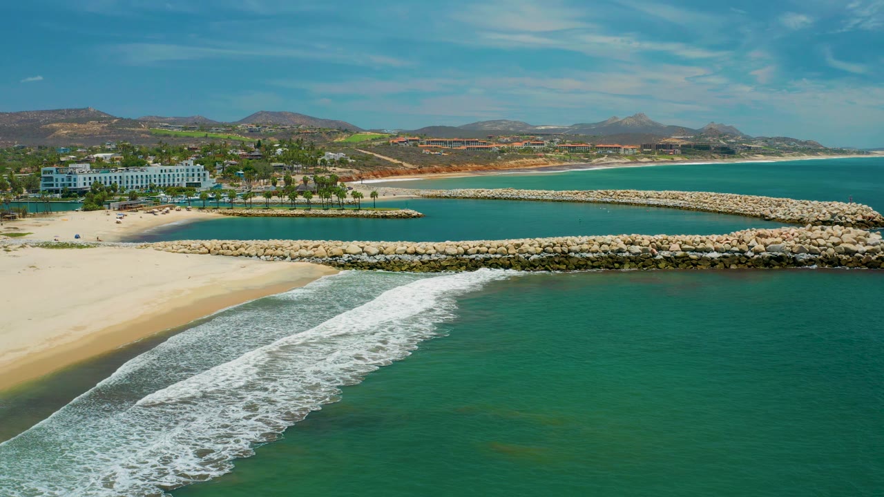 San Jose Del Cabo aerial frame showing a broad white wave band over turquoise water, pale sand at left, white rocks dividing the inlet from the sea, resort buildings, hills, and no visible people.