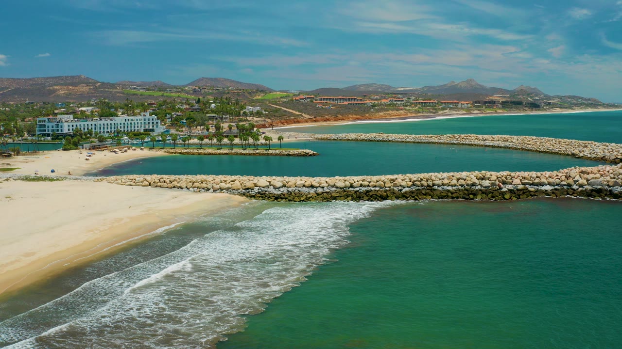 Aerial frame over San Jose del Cabo showing foamy surf in the foreground, pale beach at left, rock breakwaters across the water, resort buildings and palms beyond, and no clearly visible people.