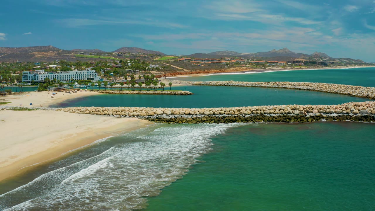 San Jose del Cabo aerial frame showing white wave foam cutting diagonally through the foreground, broad sand at left, parallel rock jetties, low resort buildings, palms, and dry hills under clear light.