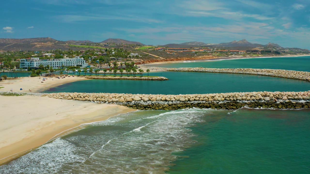 Opening aerial frame in San Jose Del Cabo, Mexico with pale sand, foamy shore break, stone breakwaters, turquoise water, a white resort building, palms, low hills, and no visible people.