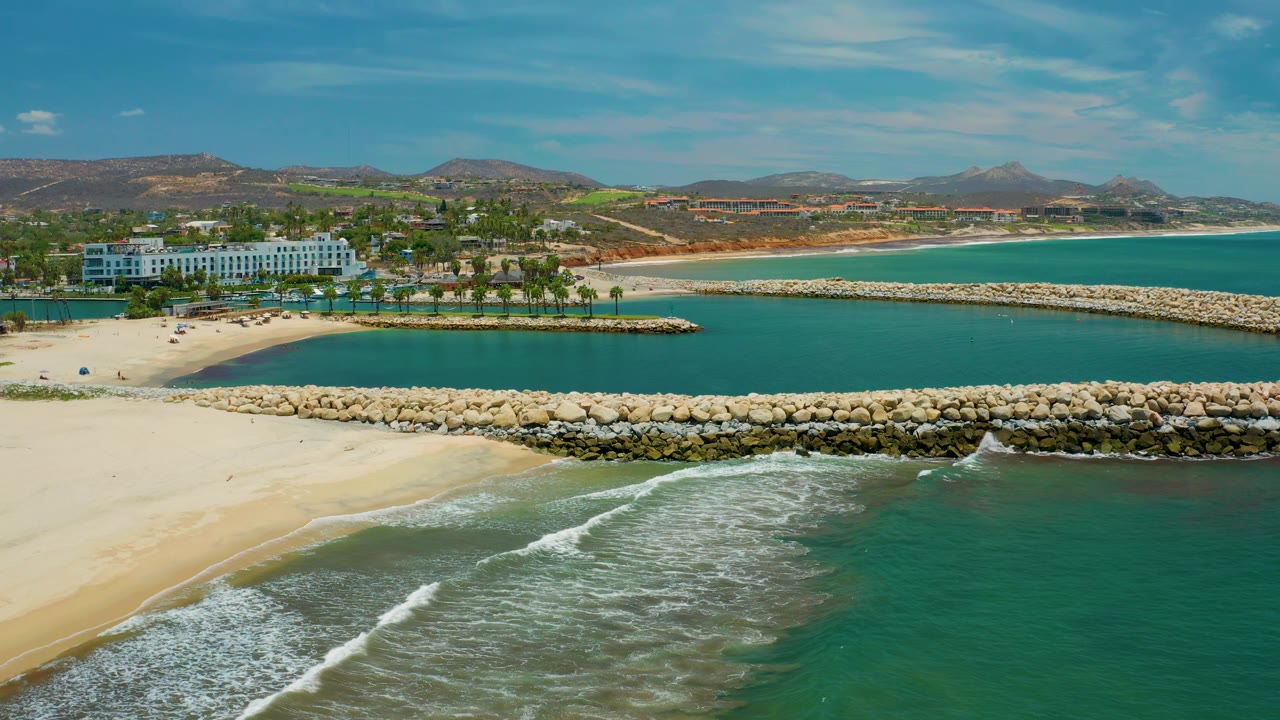 Aerial frame in San Jose Del Cabo, Mexico with white foam curling farther along the foreground beach, green water at right, two stone barriers through the center, resort buildings, palms, hills, and no visible people.