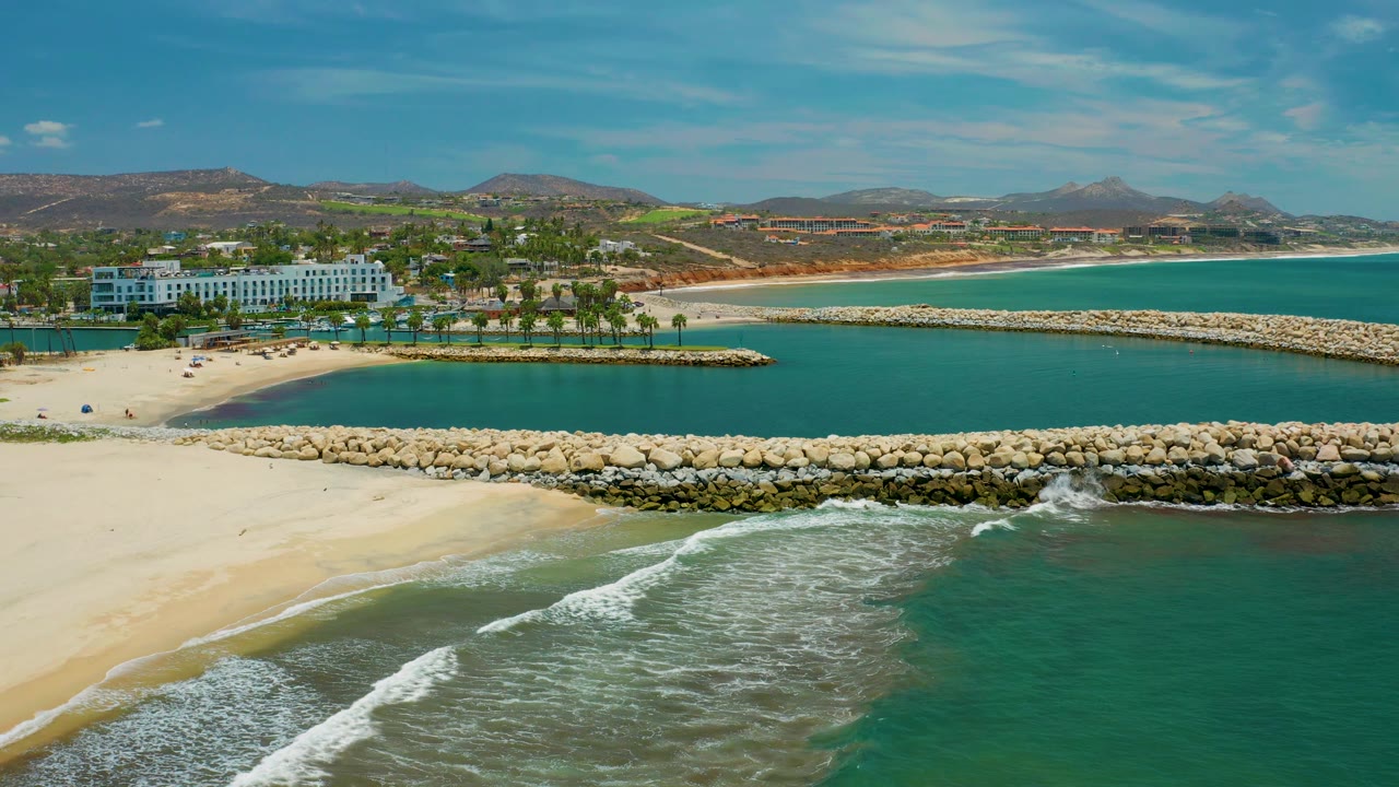 Mid-clip aerial frame in San Jose Del Cabo, Mexico showing a wider foam wash in the foreground, calm turquoise water between stone breakwaters, a beachfront resort, palms, dry hills, and no visible people.