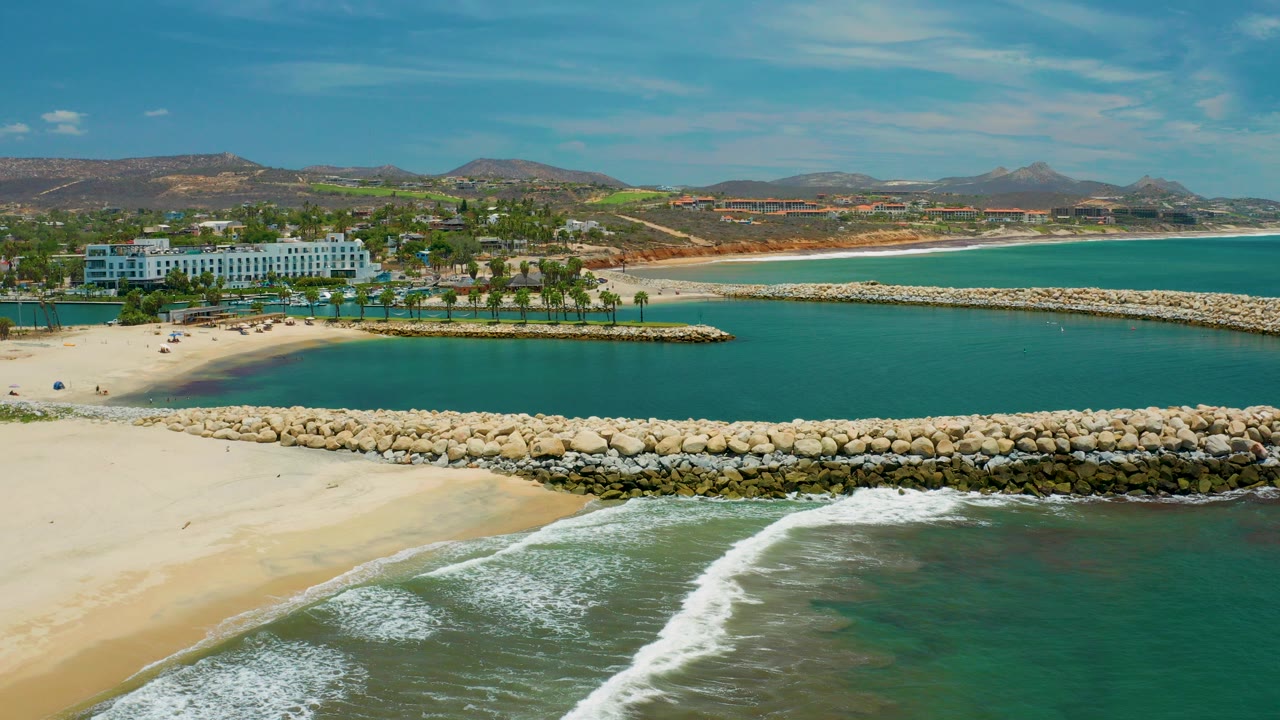 Aerial frame over San Jose Del Cabo showing surf on pale sand, a rock breakwater, blue lagoon water, a white resort, palms, distant hills, and no visible people.