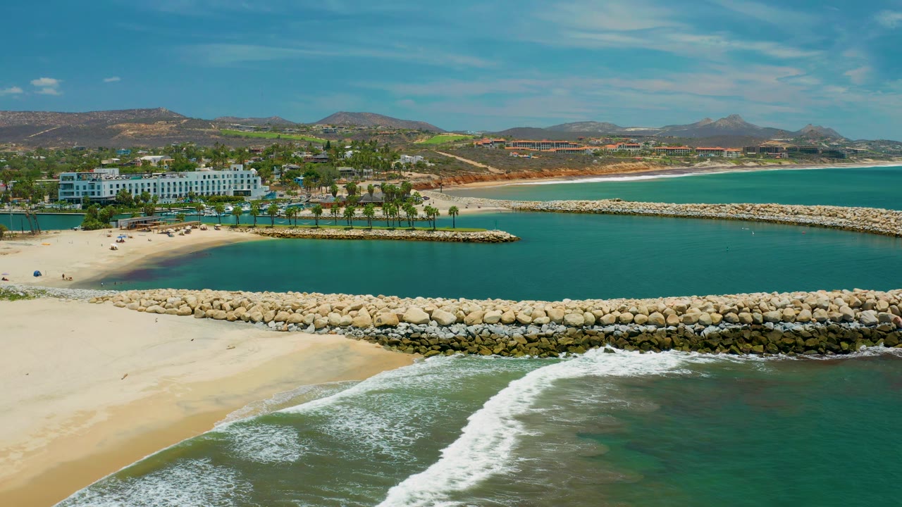 San Jose Del Cabo aerial frame showing more open water to the right, the beachfront resort and palms to the left, surf crossing the lower edge, and no visible people.