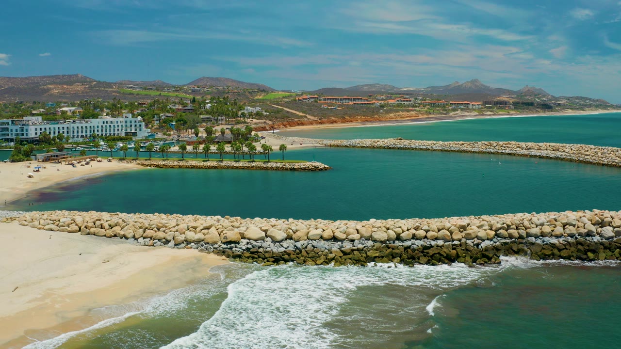 Opening aerial view in San Jose Del Cabo Mexico showing a stone breakwater across the frame, beach surf in the foreground, turquoise water, a white waterfront building, inland hills, and tiny people visible near the beach area.