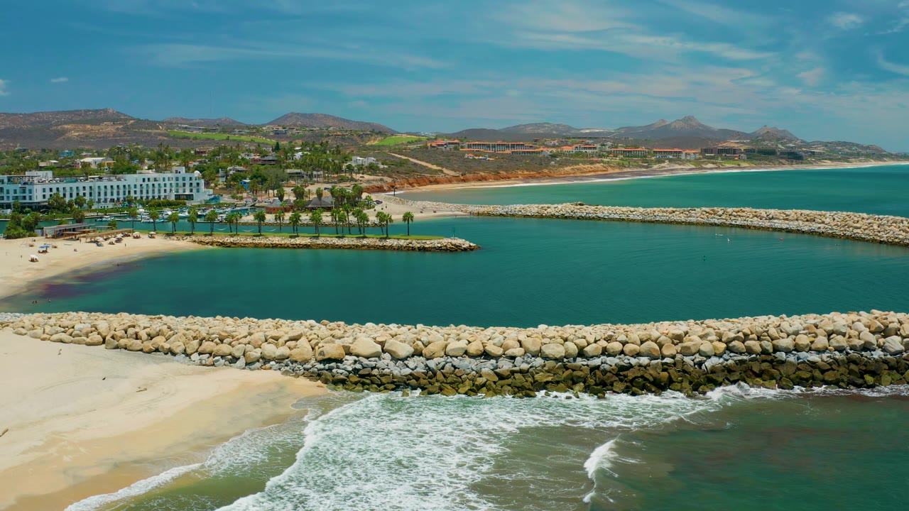 San Jose Del Cabo Mexico aerial view showing waves foaming against the sand below the stone breakwater, calm blue-green water behind it, palm landscaping, white waterfront architecture, and dry hills beyond.