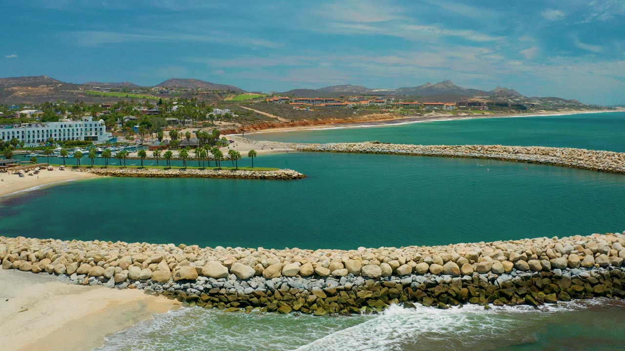 Wide aerial frame over San Jose Del Cabo coastline with turquoise water, pale rock breakwater, sandy beach, palms, white beachfront building, low hills, and no visible people.
