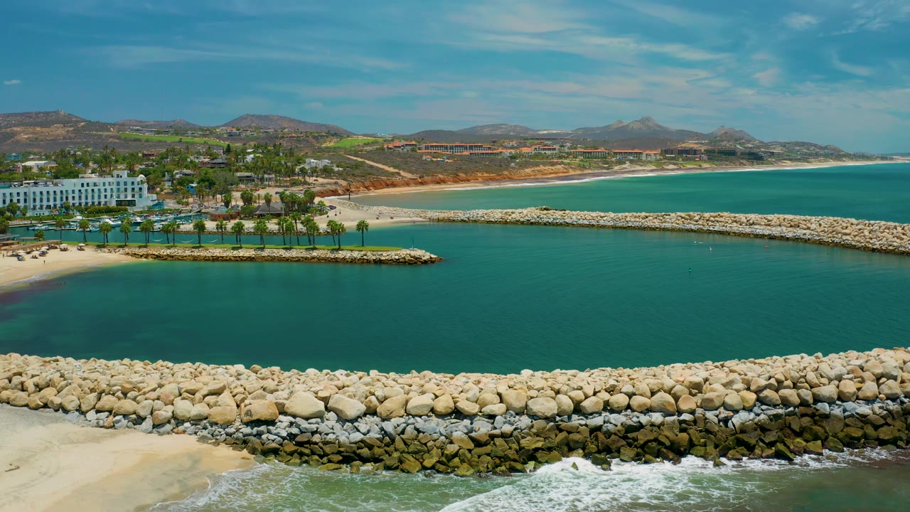 San Jose Del Cabo shoreline frame with a pale rock barrier crossing the water, a white beachfront building and palm rows to the left, dry hills beyond, and no visible people.