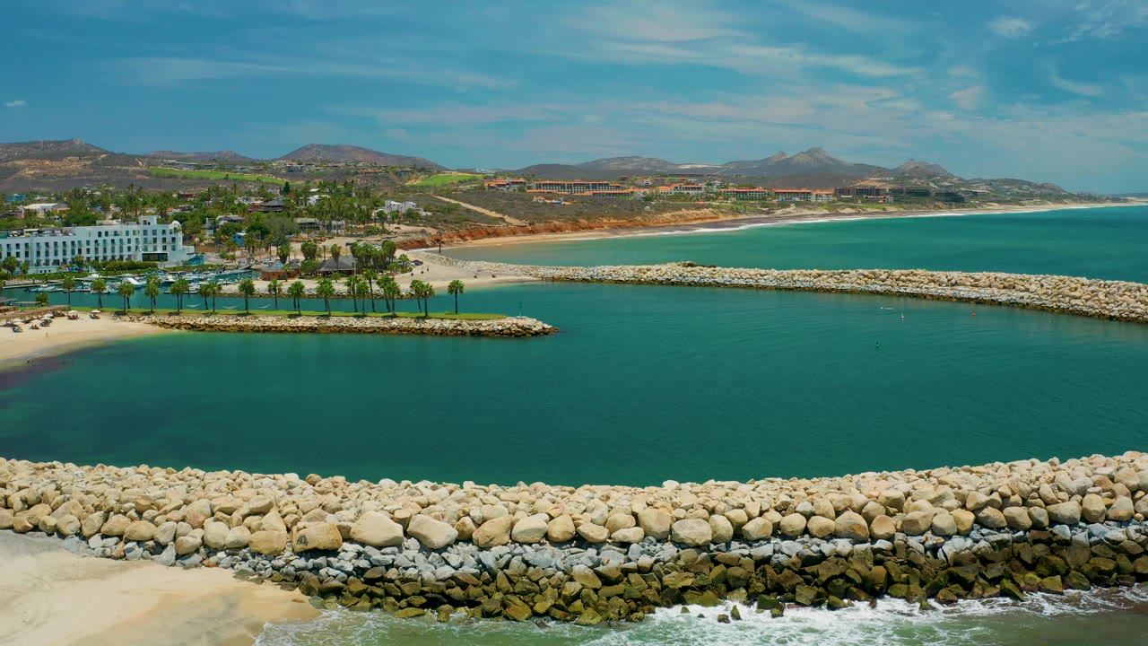 Mid-clip San Jose Del Cabo aerial frame showing turquoise water, a curving rock edge, sand, palm trees, a white building, distant hills, and no visible people.
