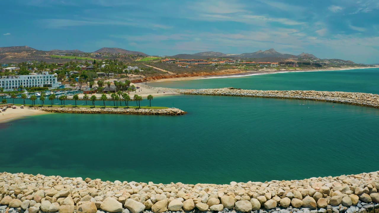 San Jose Del Cabo resort coastline seen from above, with the white hotel block left of frame, palms along the water, a curving beach, and no visible people.