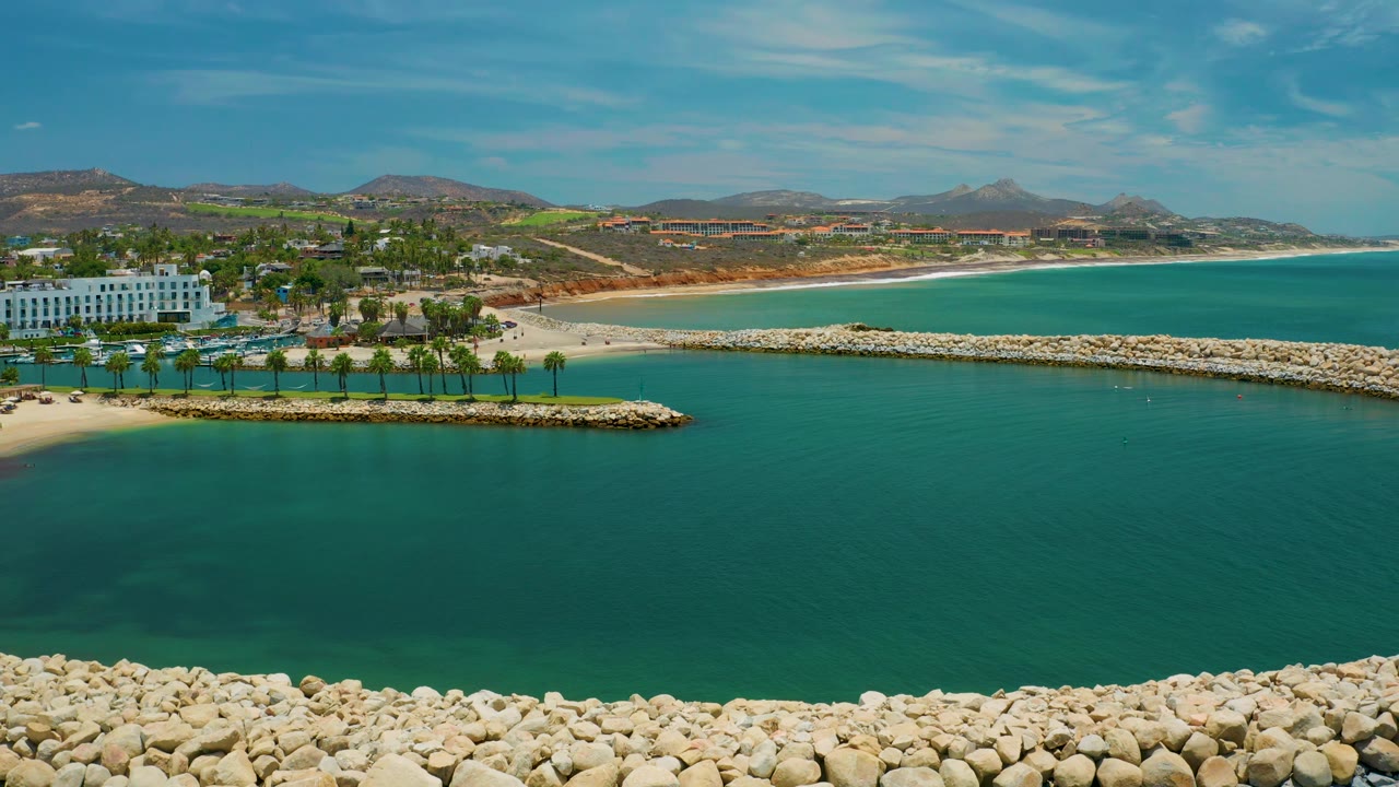 Aerial view of the San Jose Del Cabo beachfront with a long rocky breakwater crossing the frame, resort grounds at left, open blue-green water, and no visible people.