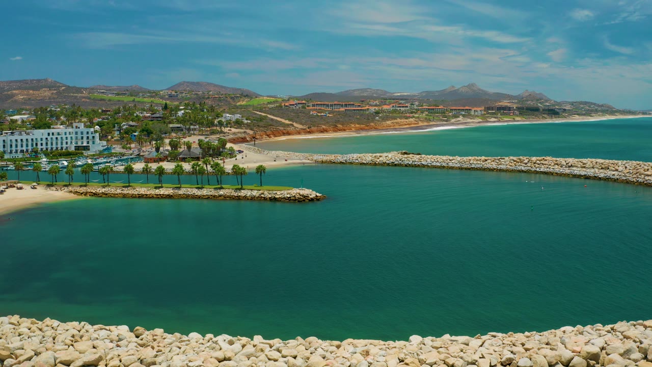 San Jose Del Cabo aerial frame with pale rocks along the foreground edge, calm turquoise water, a palm-lined resort area, sandy beach, distant hills, and no visible people.
