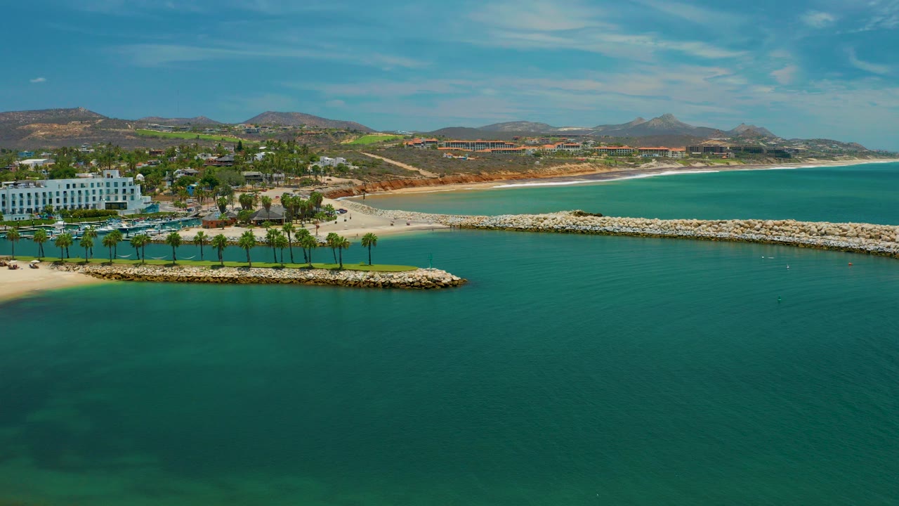 Aerial view of turquoise water, palm-lined beach, breakwater, white beachfront building, and coastal hills.