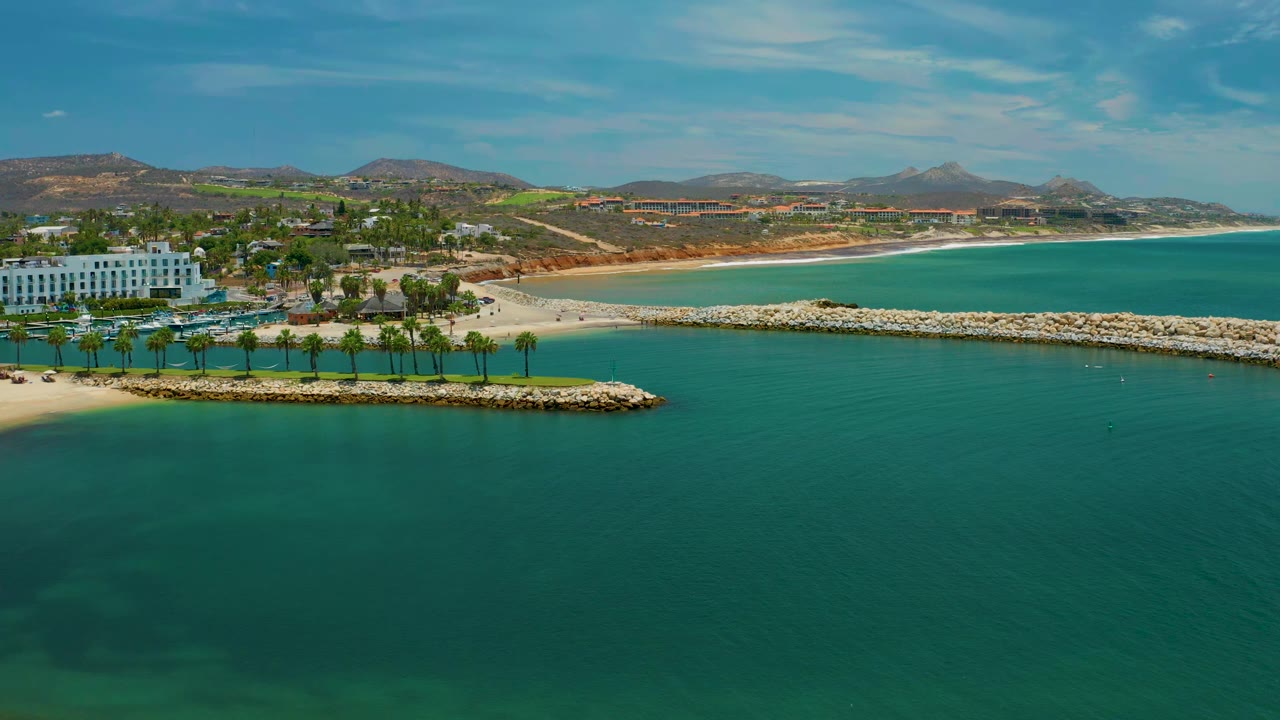 San Jose Del Cabo aerial view with teal water covering the lower frame, the rock breakwater stretching across the beach, palms near a white building, low hills, and no visible people.